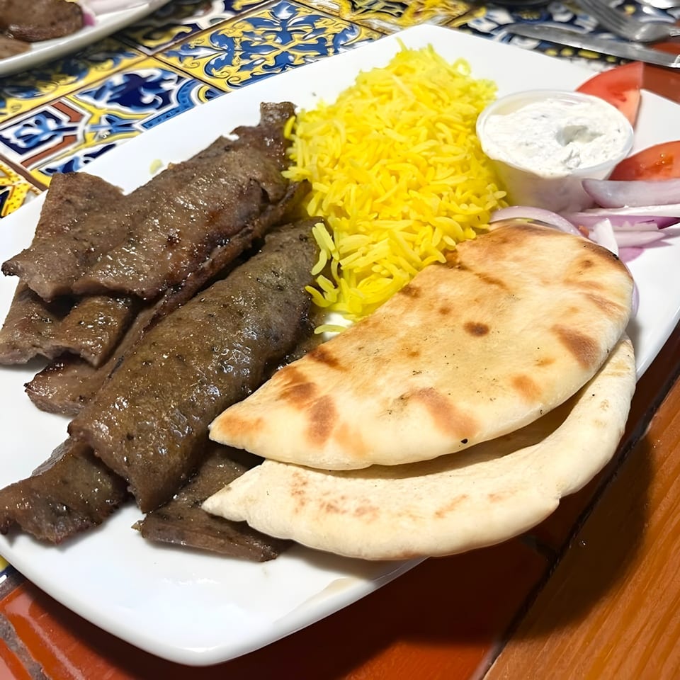 A Mediterranean gyro plate with sliced meat, yellow rice, pita bread, tzatziki, tomatoes, and onions.