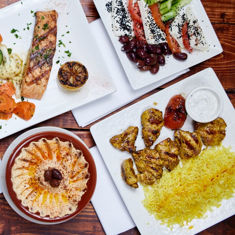 Grilled salmon, chicken skewers with rice, hummus, and a Mediterranean salad on a wooden table.
